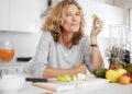 Smiling woman leans at a kitchen counter with cut fruit, a blender, citrus, pineapple, bananas, and juice.